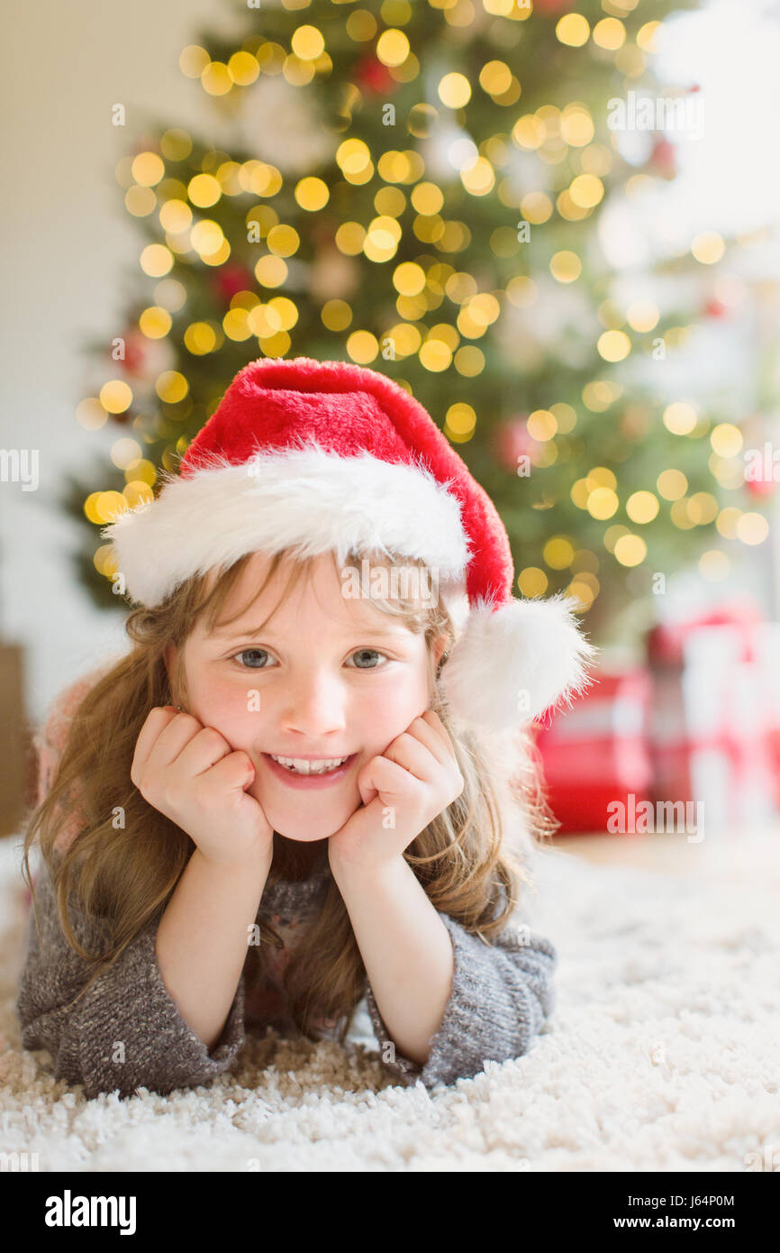 Ritratto ragazza sorridente indossando Santa hat sul tappeto nel soggiorno con albero di Natale Foto Stock