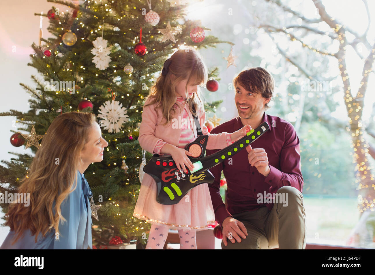 I genitori a guardare la figlia suonare la chitarra giocattolo regalo di Natale nella parte anteriore dell albero di Natale Foto Stock