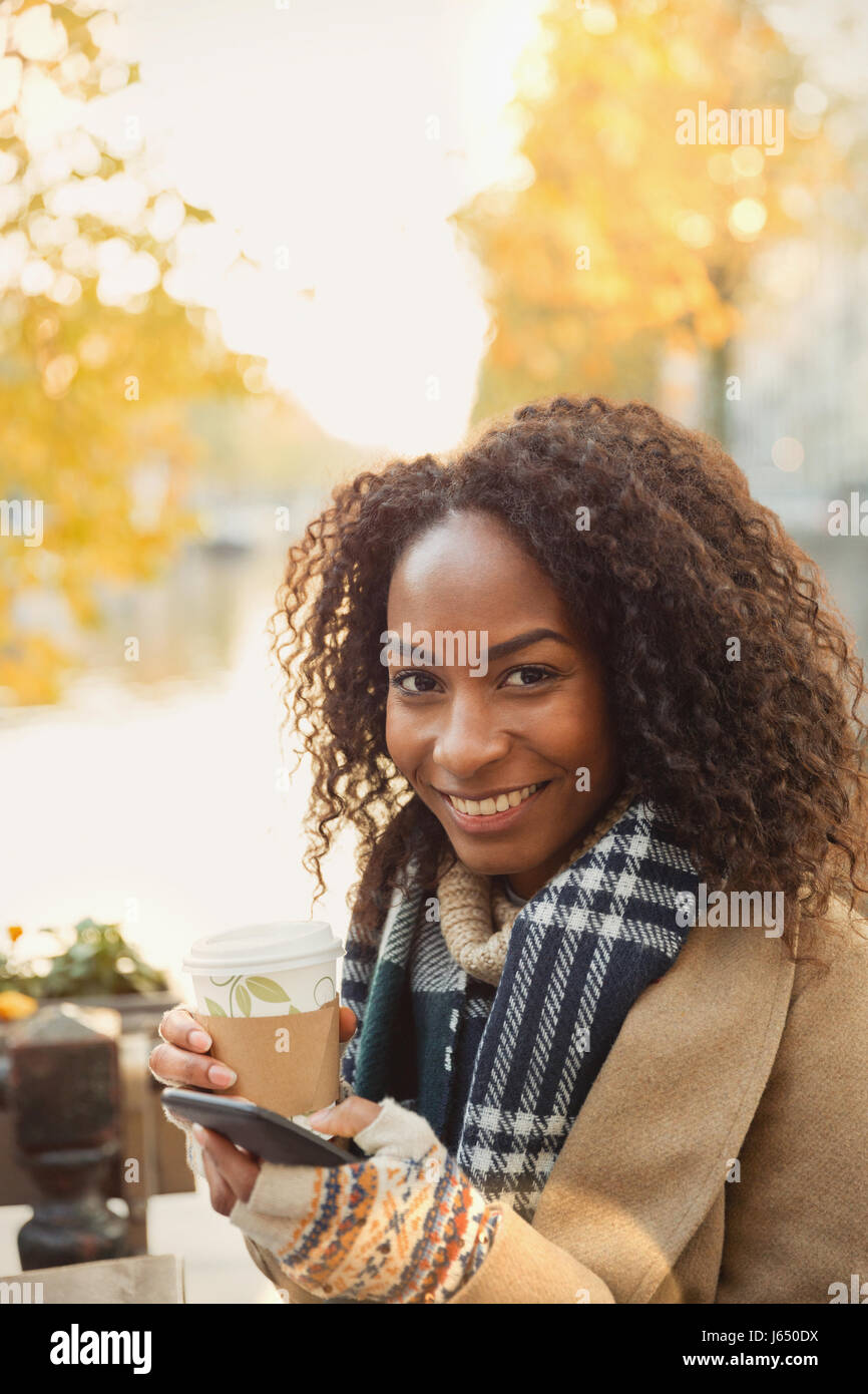 Ritratto sorridente giovane donna di bere il caffè e i messaggi di testo con un telefono cellulare a livello urbano sidewalk cafe Foto Stock