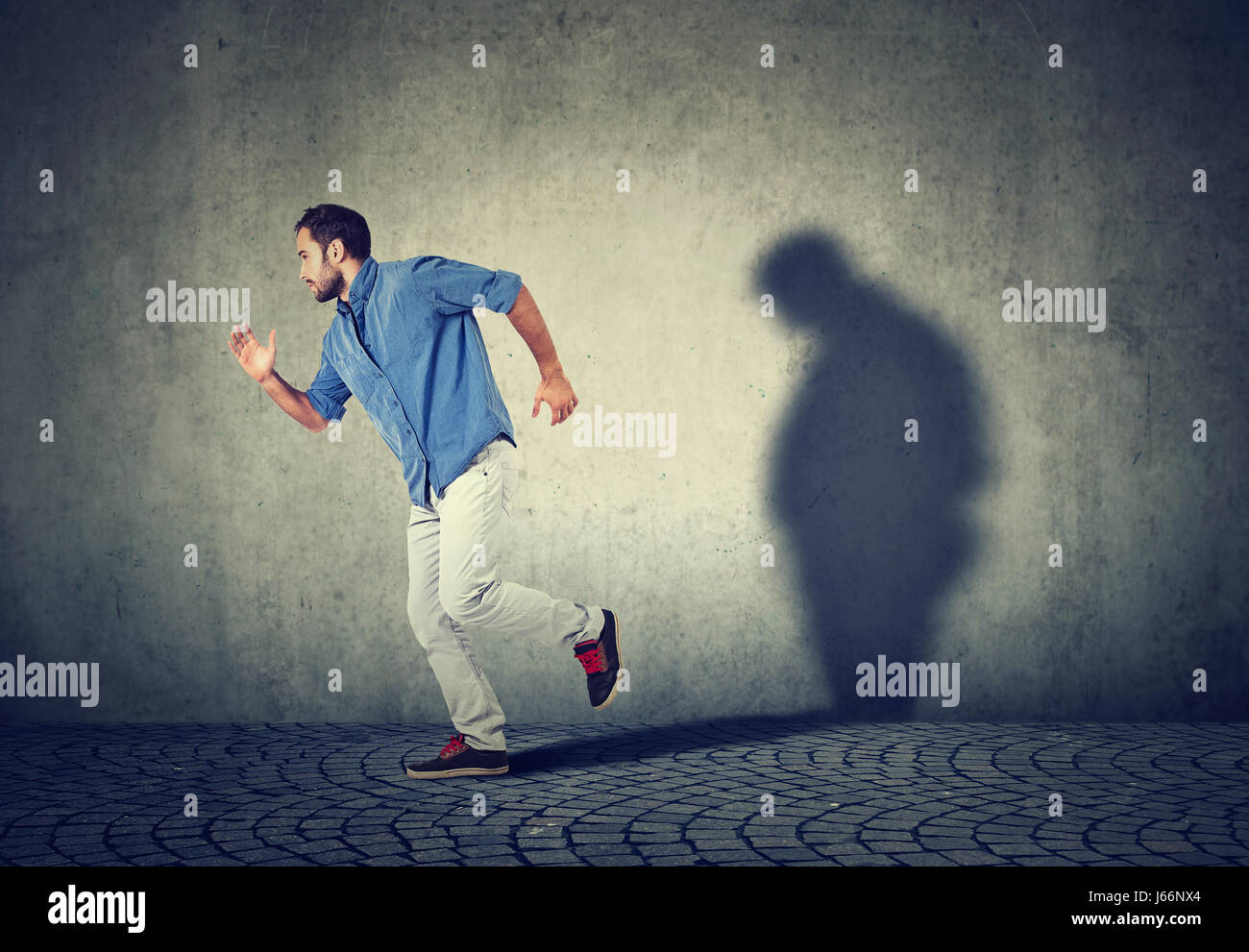 Uomo che corre lontano dal suo triste cupo fat ombra sul muro. Salute mentale e controllo del peso corporeo concept Foto Stock