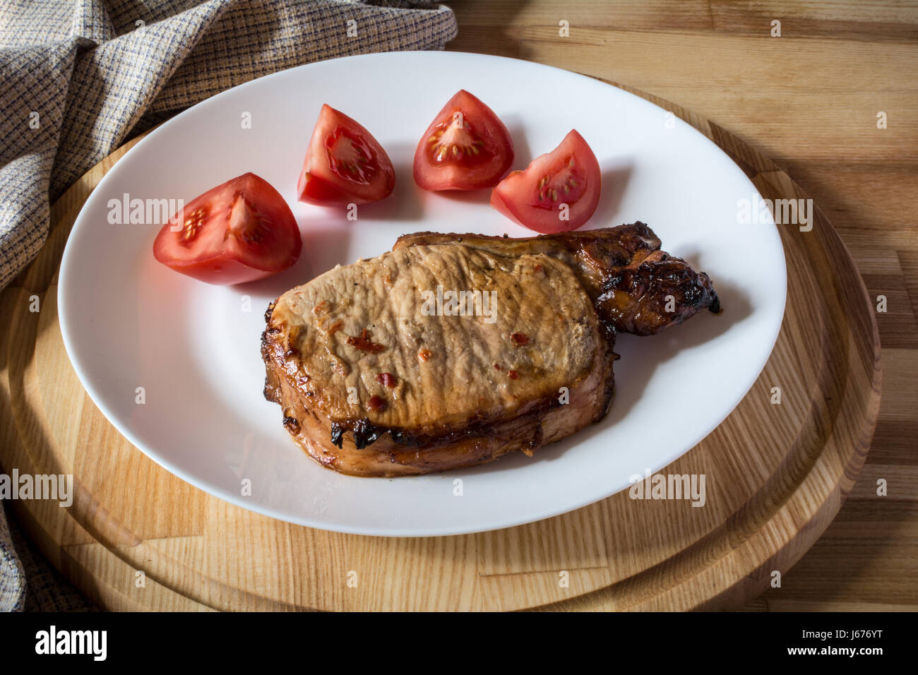 Bistecca di carne di maiale e fette di pomodoro su una piastra bianca su una tavola di legno vassoio rotondo Foto Stock