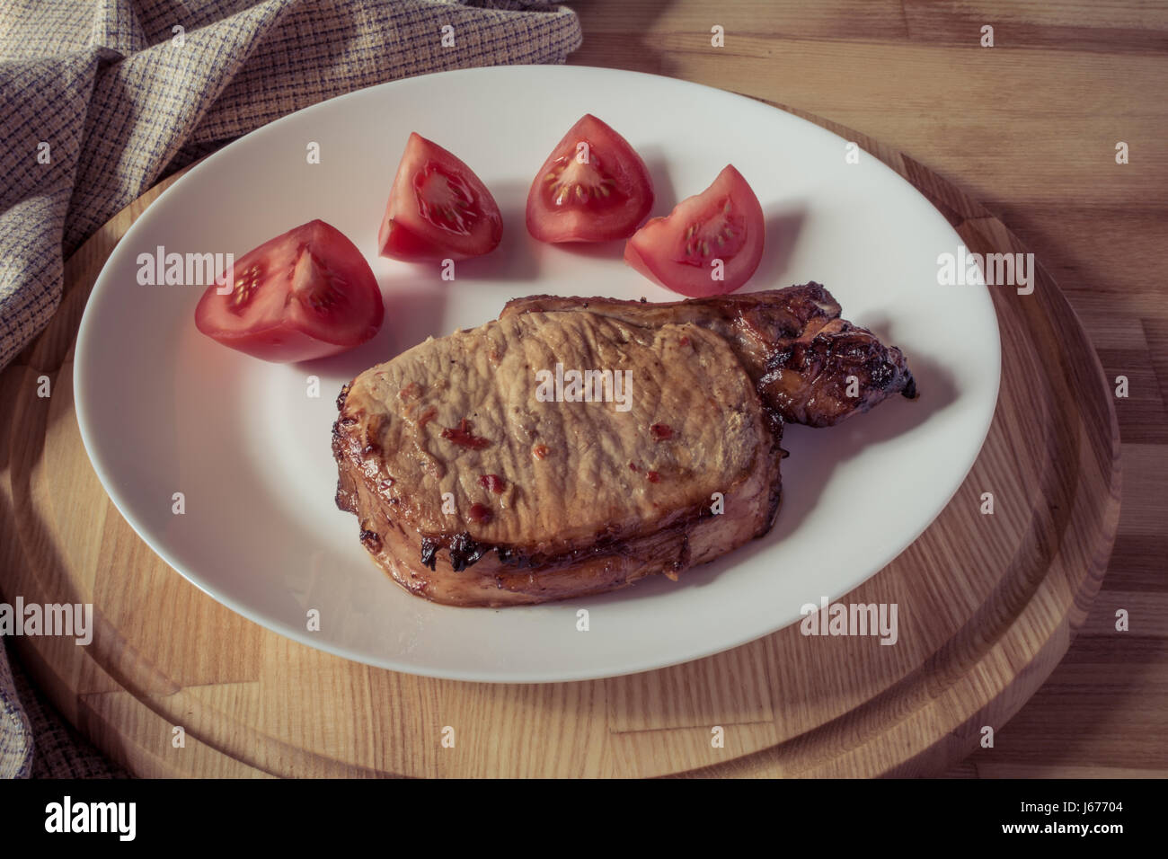 Bistecca di carne di maiale e fette di pomodoro su una piastra bianca su una tavola di legno vassoio rotondo, effetto vintage Foto Stock