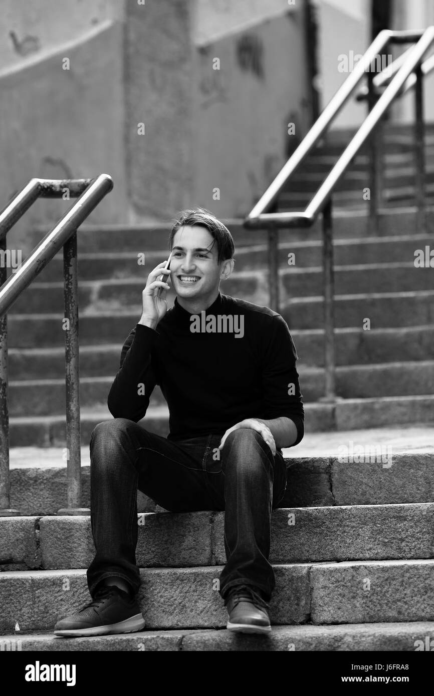 Giovane uomo parlando sul cellulare durante la seduta sulle scale all'esterno. In bianco e nero photo. Foto Stock