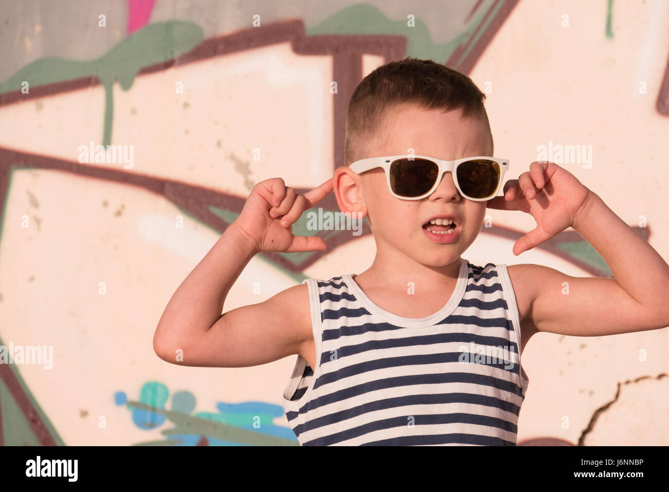 Fantastico piccolo bambino maschio bianco da indossare occhiali da sole e  maglietta a strisce sul muro di graffiti sullo sfondo di toccare le sue  orecchie con le mani Foto stock - Alamy