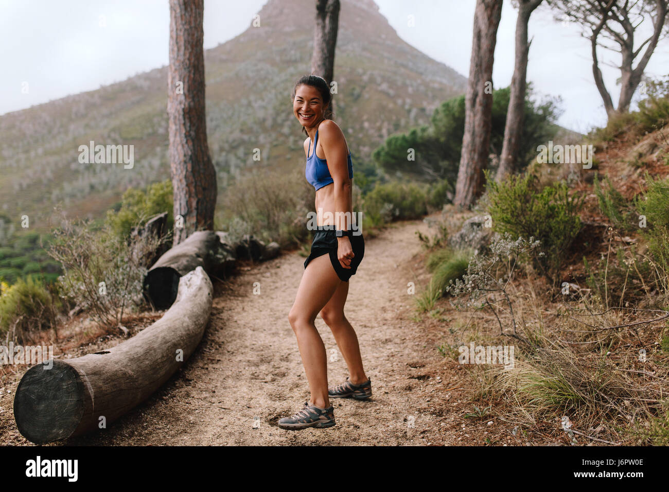 A piena lunghezza shot di felice giovane donna asiatica in piedi sul sentiero di montagna e sorridente. Donna Fitness in sportswear su cross country percorso. Foto Stock