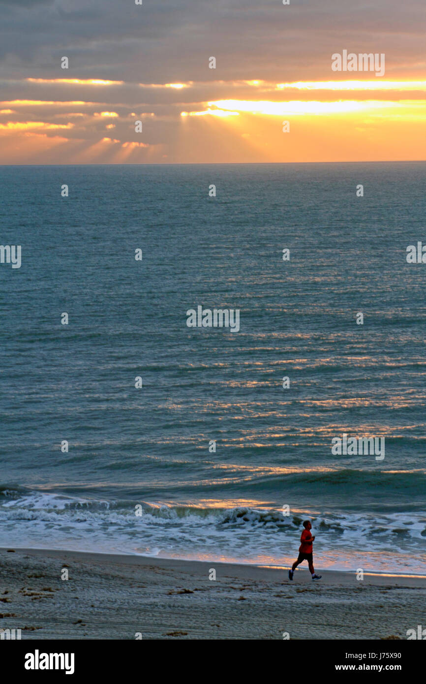 Golden raggi di luce rompere attraverso il buio clounds per versare su un mare scintillante di un uomo solitario corre lungo la spiaggia Foto Stock