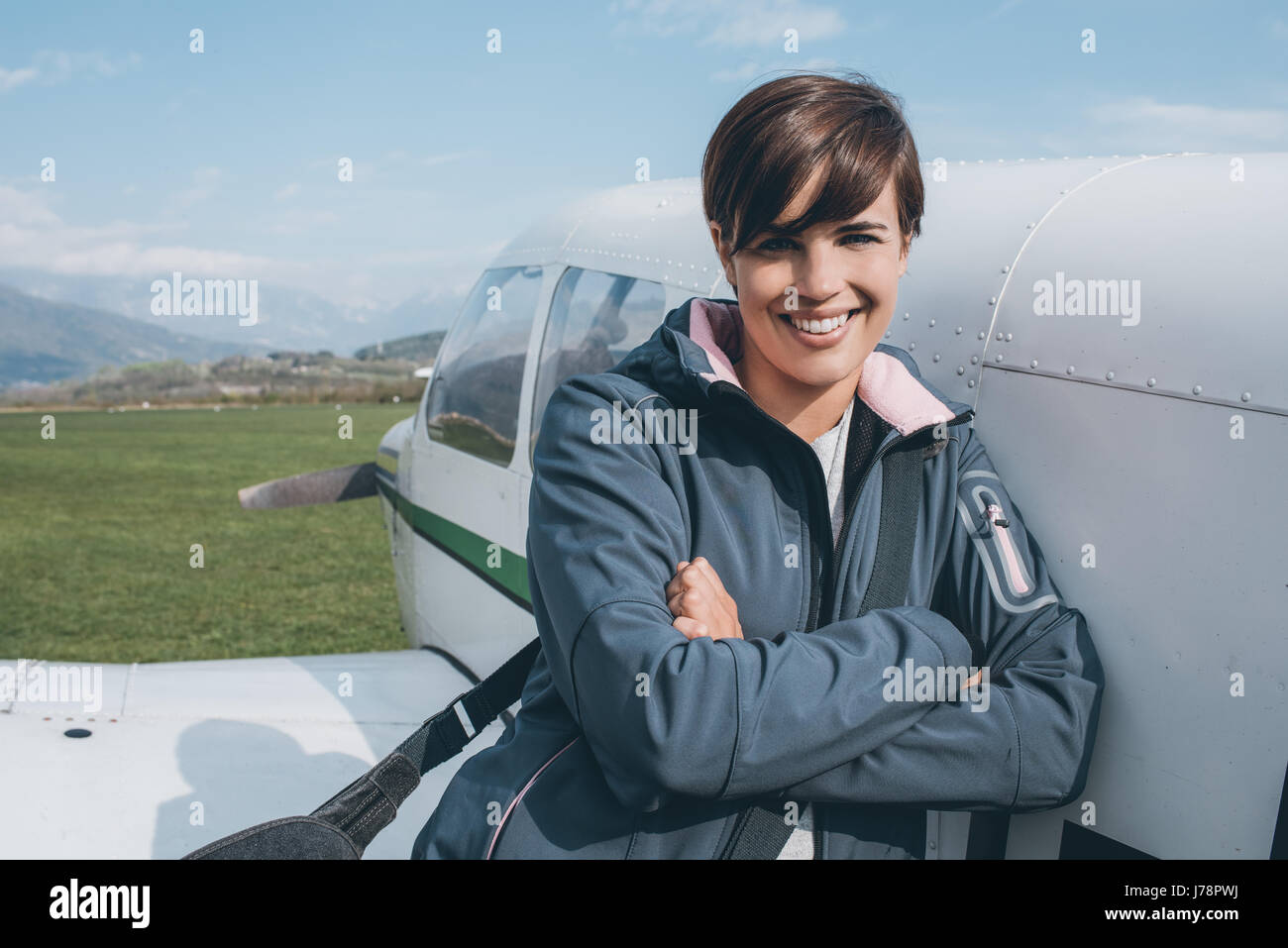 Sorridendo allegro pilota femmina appoggiata su un aeroplano ad elica e posa, cielo blu sullo sfondo, viaggi e concetto di aviazione Foto Stock