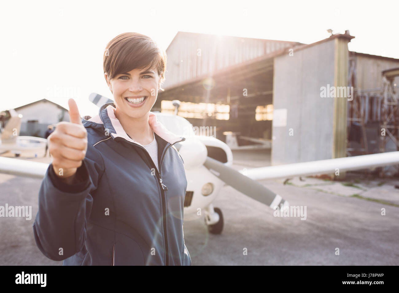 Sorridente giovane pilota che pongono in aeroporto e dando un pollice in alto, elica di piano sullo sfondo, viaggi e concetto di aviazione Foto Stock