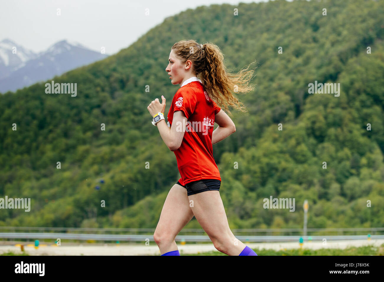 Redhead girl runner in esecuzione sullo sfondo delle montagne e alberi verdi in gara Spring Mountain marathon Foto Stock