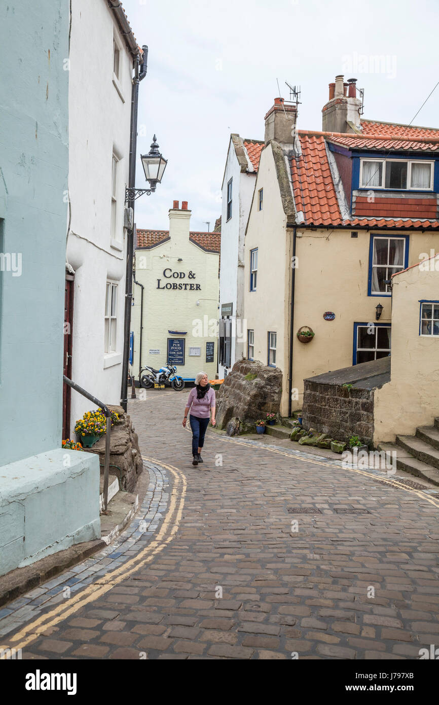 Una donna cammina lungo le strade acciottolate e tortuose di Staithes, North Yorkshire, Regno Unito Foto Stock