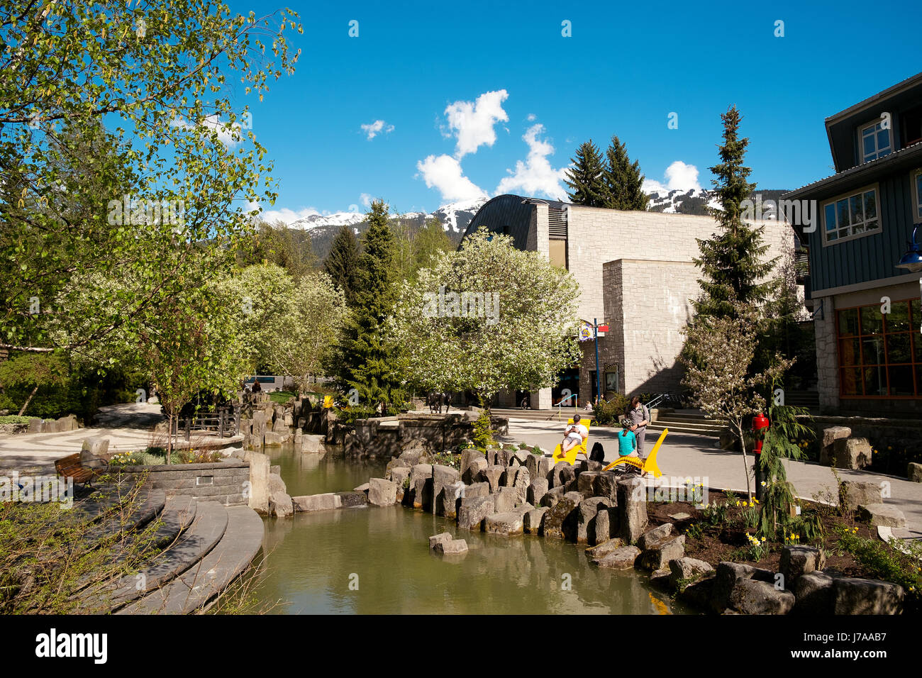 Il Whistler in una giornata di sole in primavera. Persone sedersi dal villaggio di Whistler corso d'acqua che scorre accanto al Maury giovani Arts Centre Whistler BC, Foto Stock