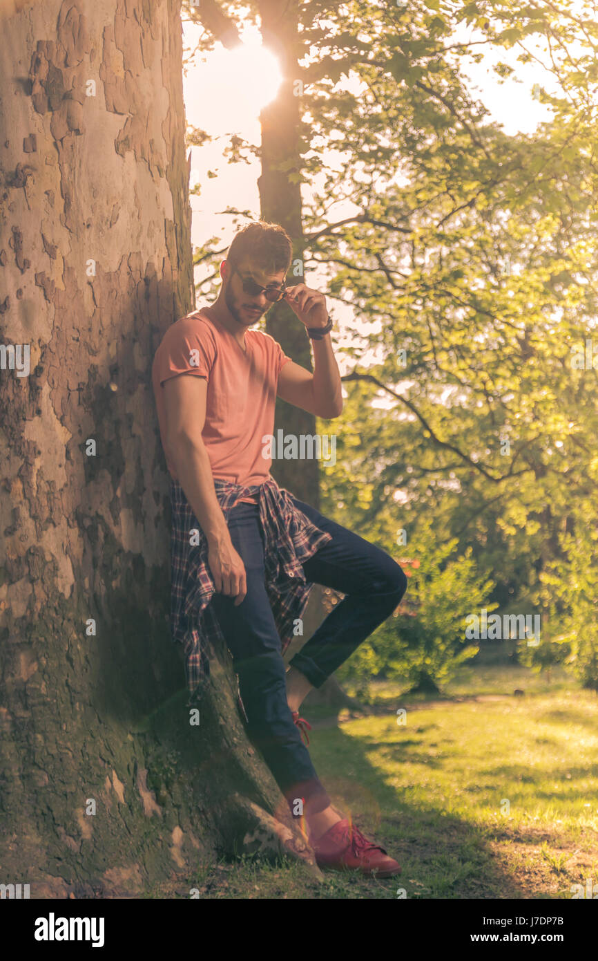 Un giovane uomo adulto, parco natura all'aperto, poggiando su albero, giornata soleggiata, abiti casual, Foto Stock