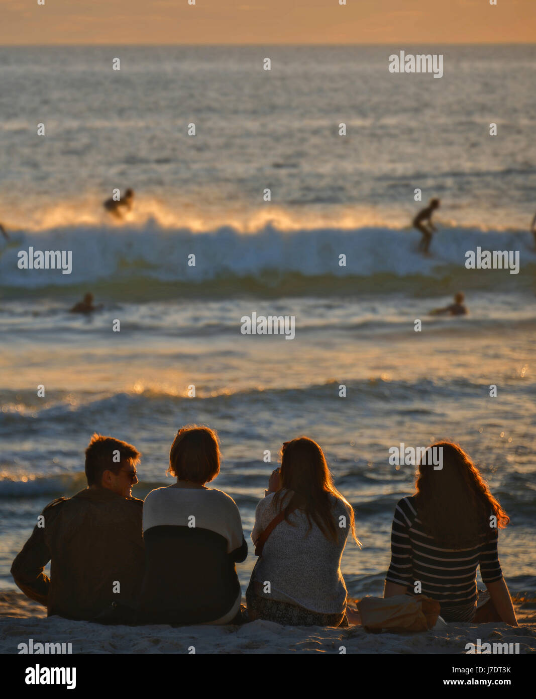 Un'immagine della vista posteriore di quattro persone in primo piano al tramonto, con Surfers cattura onde in background. Cottesloe Beach, Australia occidentale Foto Stock