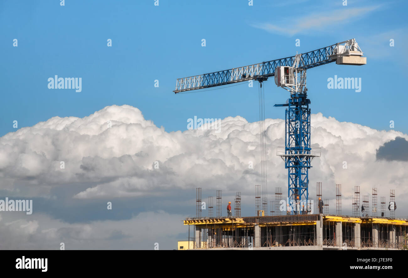 La costruzione di gru a torre è blu accanto ai costruttori sulla parte superiore di un edificio in costruzione. Su uno sfondo di nuvole e cielo blu Foto Stock