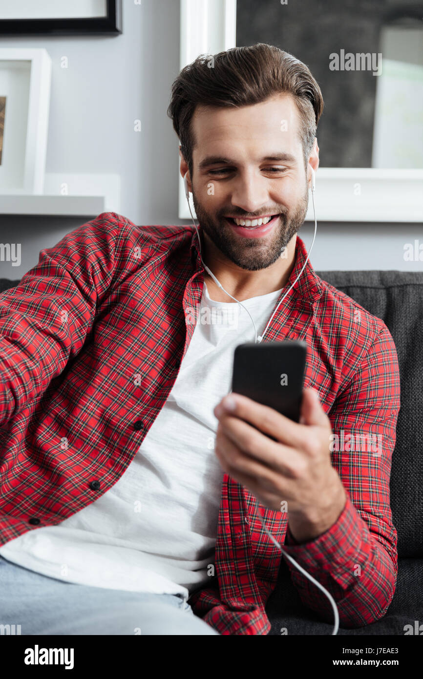 Giovane uomo barbuto in auricolari utilizza lo smartphone per effettuare una videocall Foto Stock