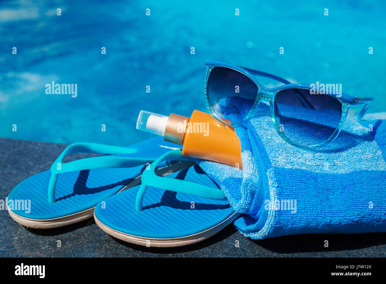 Occhiali da sole con crema di protezione solare, blu pantofole e asciugamano sul bordo di una piscina - vacanza concetto tropicale Foto Stock
