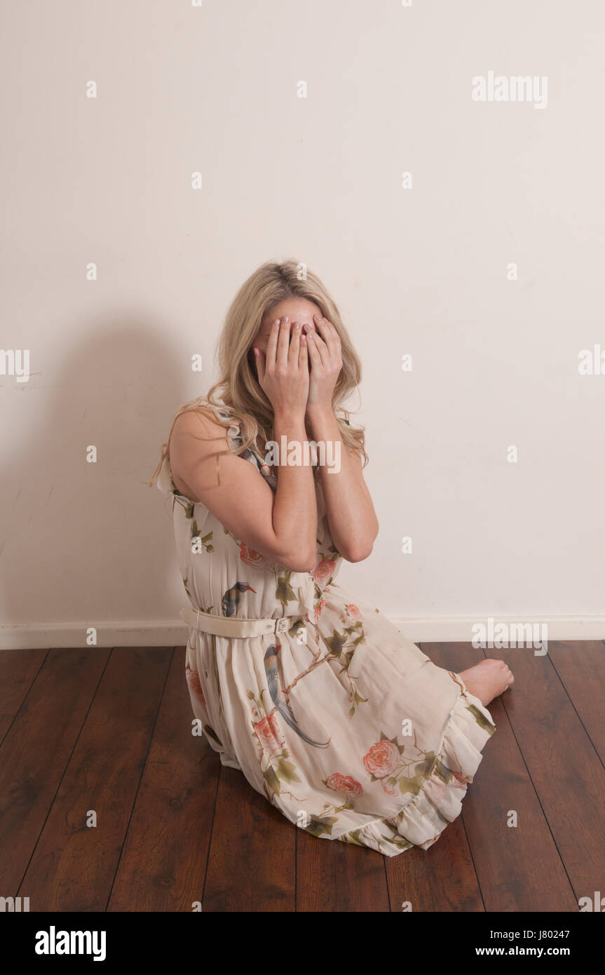 Spaventata donna bionda che nasconde il viso con le mani seduta sul pavimento a piangere Foto Stock