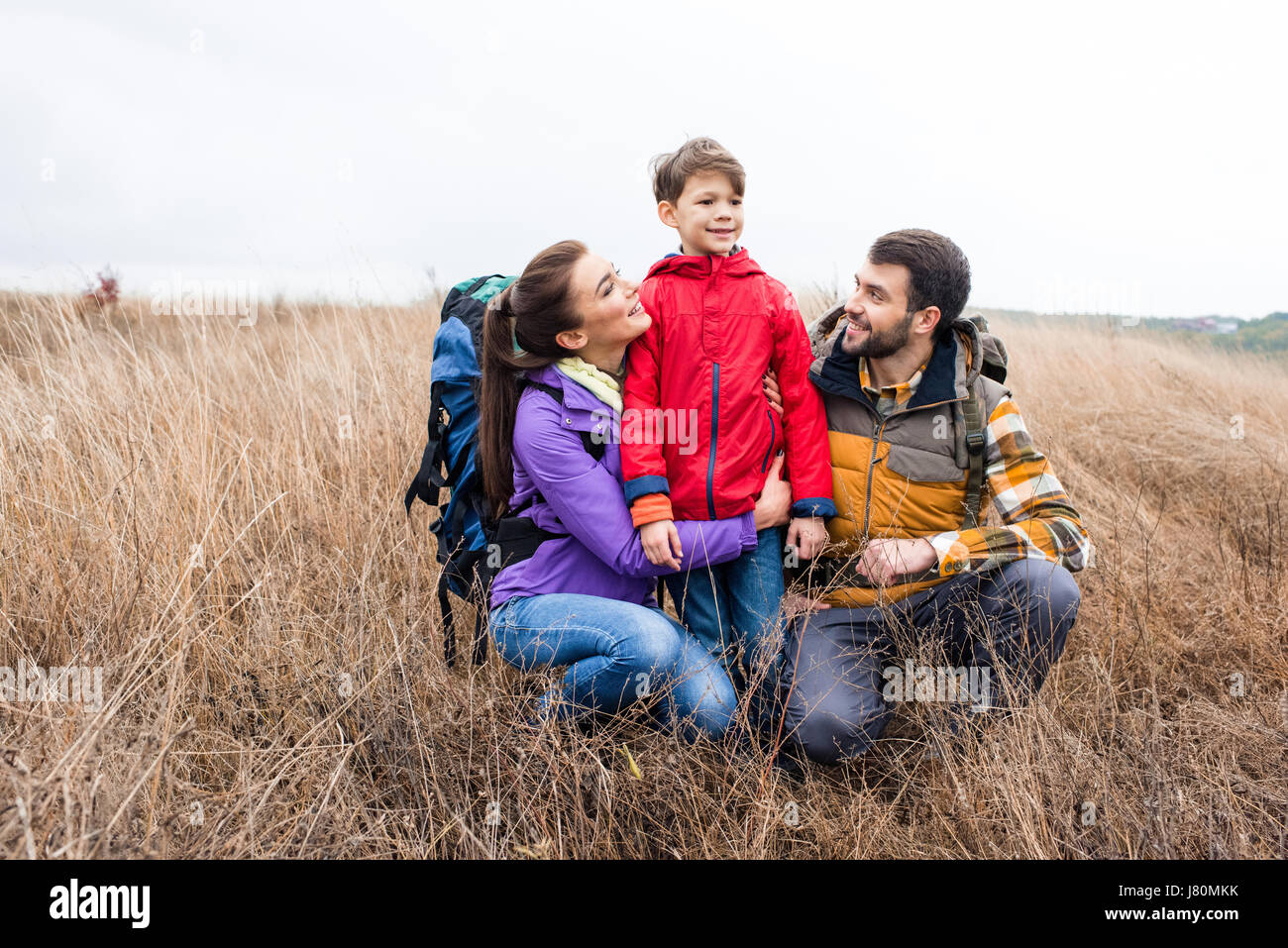 La famiglia felice con zaini abbracciando in alti erba secca e guardando lontano a nuvoloso giorno di autunno Foto Stock