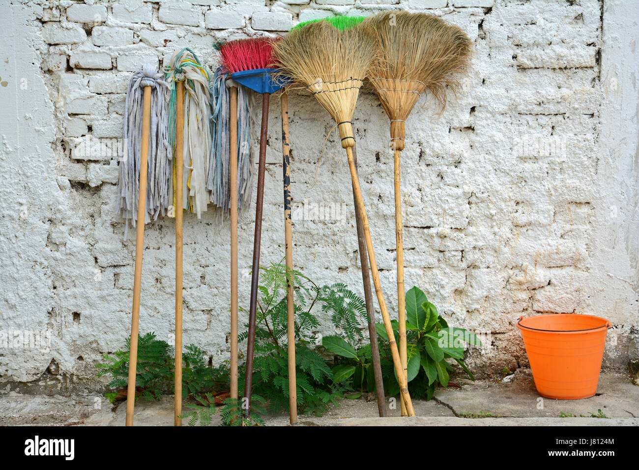 Spazzoloni di vecchio stile, scope di mais e scope di paglia appoggiate su un muro di mattoni bianchi e un secchio arancione. Foto Stock