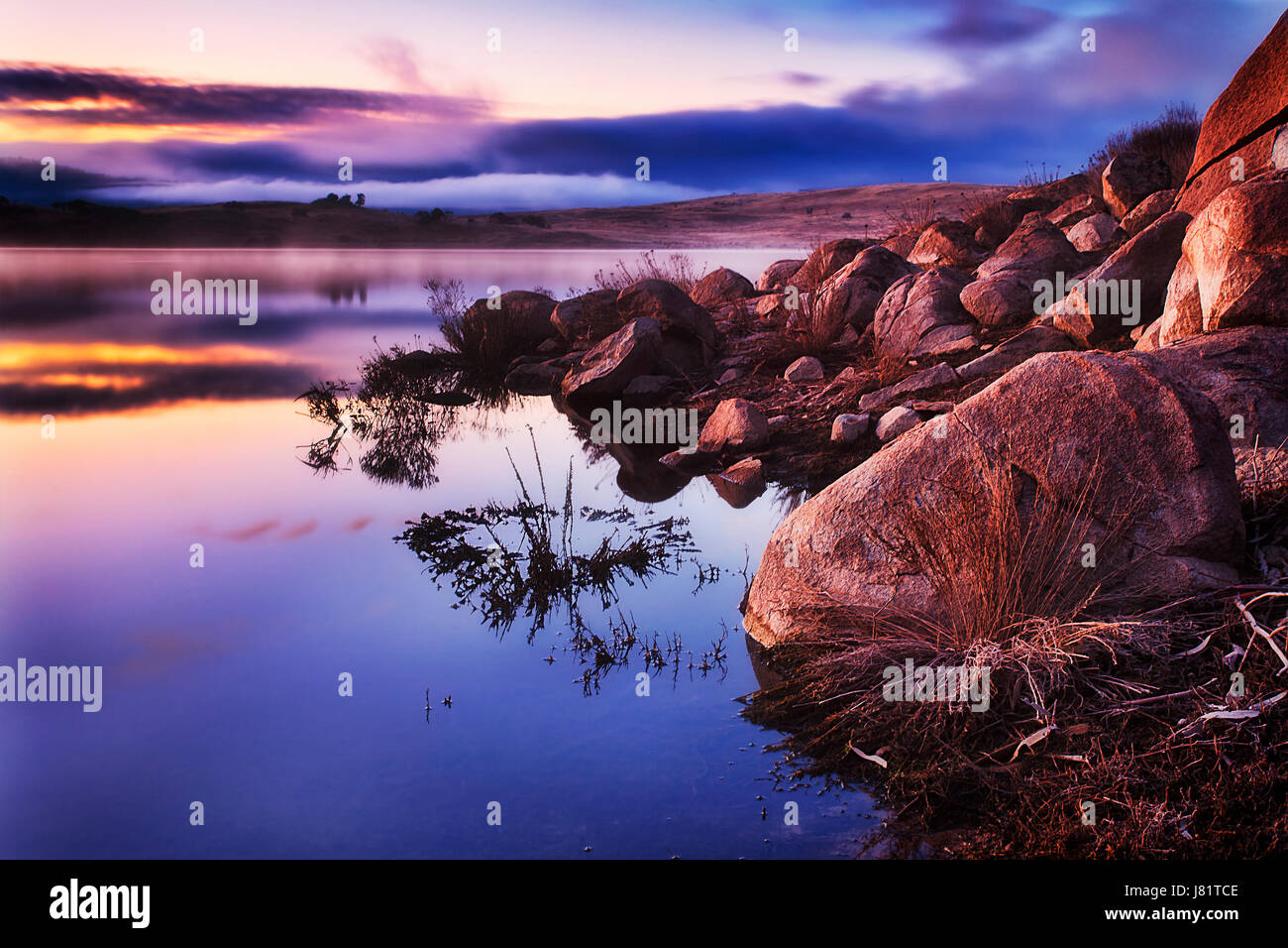 Colorato alba sul lago Jindabyne nelle montagne innevate regione del NSW, Australia. Rocce di arenaria e massi sulla riva del lago illuminato dal sorgere del sole in inverno Foto Stock