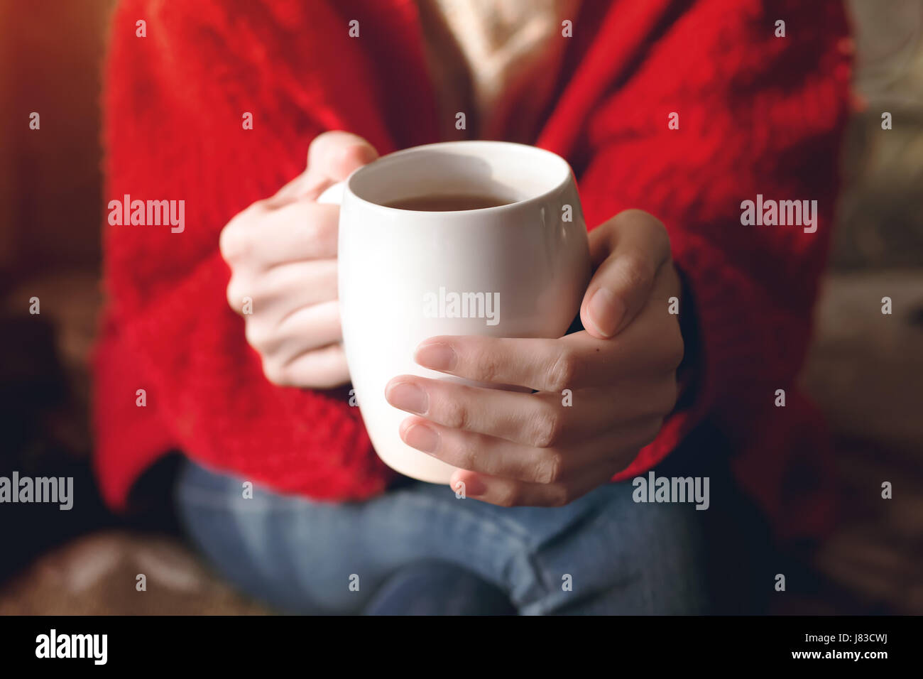Primo piano delle mani femminili con una tazza di bevanda. Bella ragazza in maglione rosso coppa di ritegno del tè nella luce del sole di mattina. Foto Stock