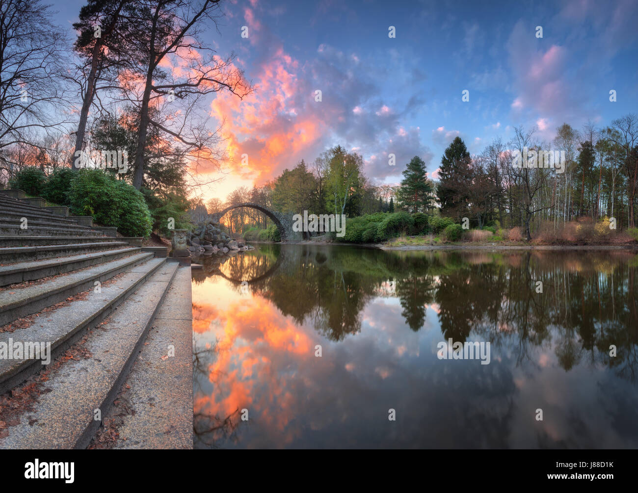 Ponte del diavolo in Kromlau, Germania. Un paesaggio fantastico con ponte Rakotz, scale, lago, alberi verdi e colorati di cielo blu con nuvole multicolori reflec Foto Stock