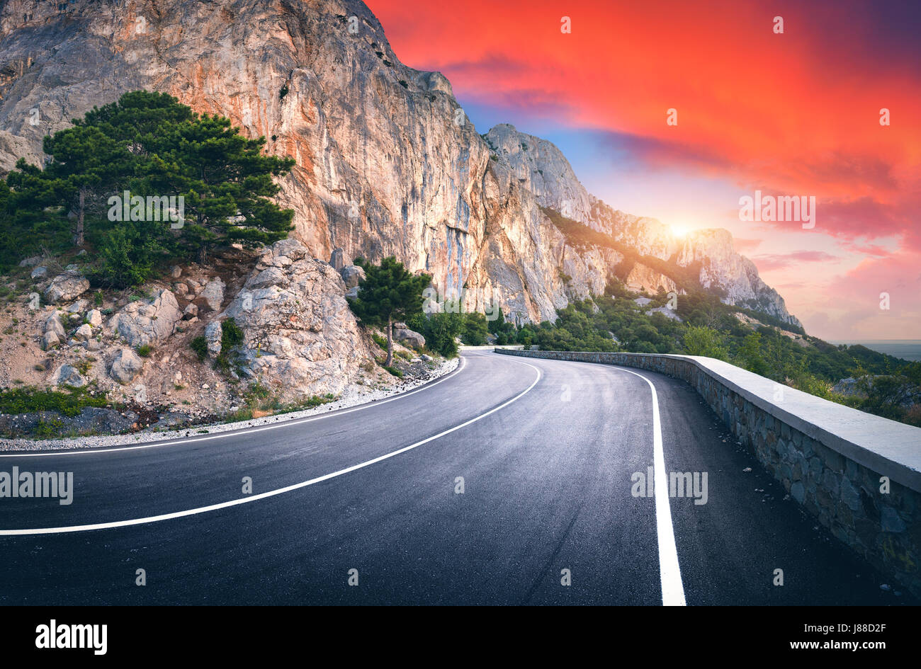 Strada asfaltata. Paesaggio con una bella strada di montagna con un asfalto perfetto, alte rocce, alberi, incredibile cielo rosso con le nuvole al tramonto in summe Foto Stock