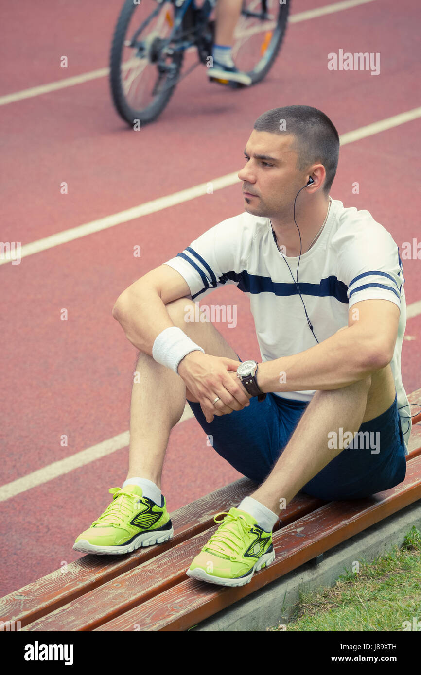 Sportivo in appoggio sul banco di prova dopo il suo allenamento mattutino. Egli è il relax ascoltando la sua musica preferita. Rosso acceso le vie e il ciclista in background Foto Stock