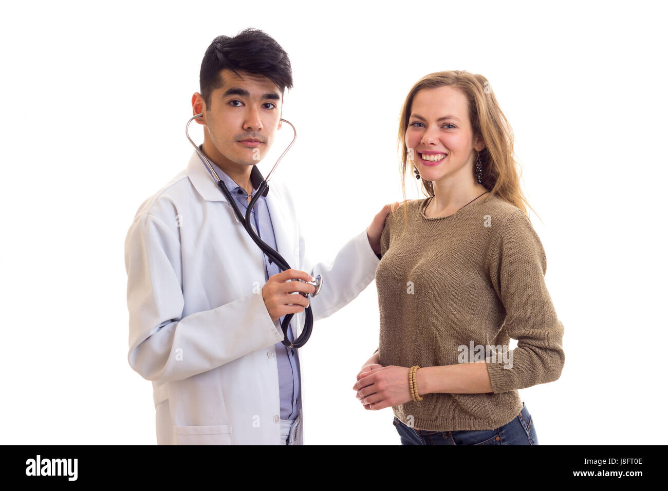 Bel giovane medico con i capelli scuri in abito bianco con uno stetoscopio ascoltando il battito del cuore della giovane donna sorridente con capelli lunghi marrone a MAGLIONE Foto Stock