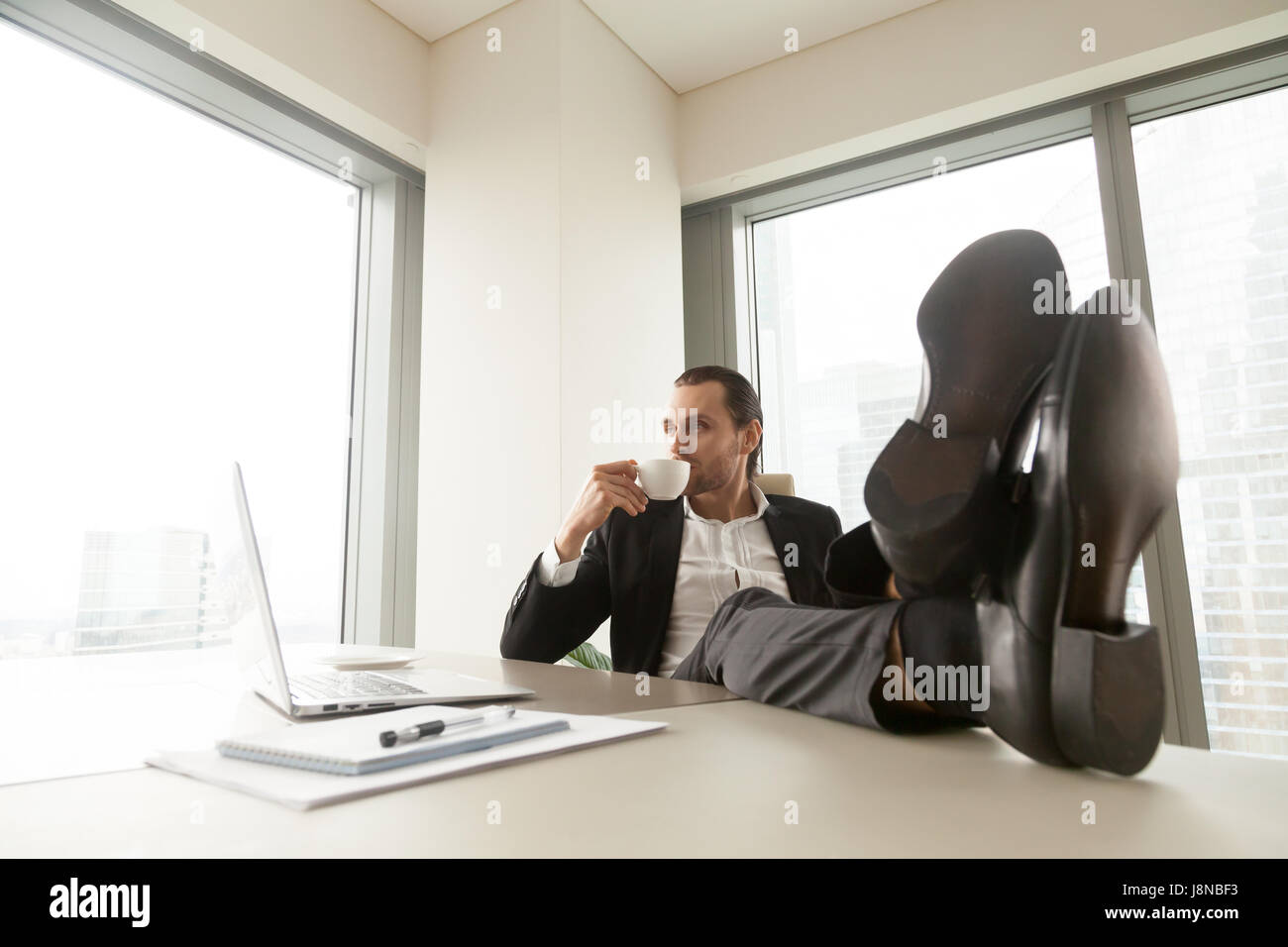 Imprenditore rilassato con le gambe sulla scrivania beve caffè in ufficio. Imprenditore di successo in appoggio al lavoro dopo una buona giornata di lavoro. Azienda leader tak Foto Stock
