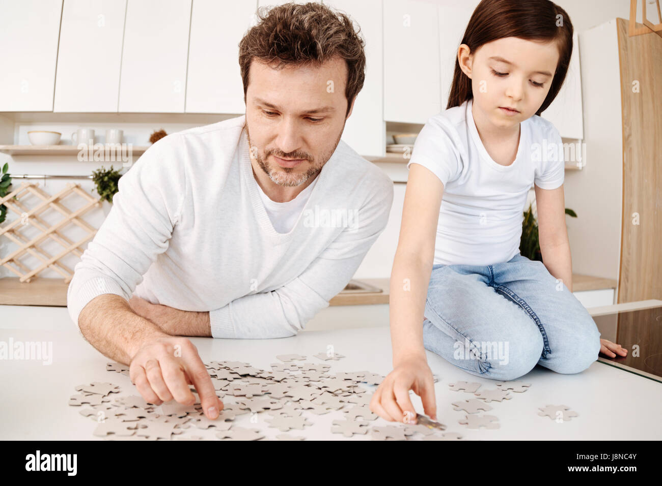 Affascinato da puzzle. Delicato Pretty girl seduto sulle sue ginocchia sul bancone della cucina e facendo un puzzle insieme con il suo padre, essendo entrambi Foto Stock