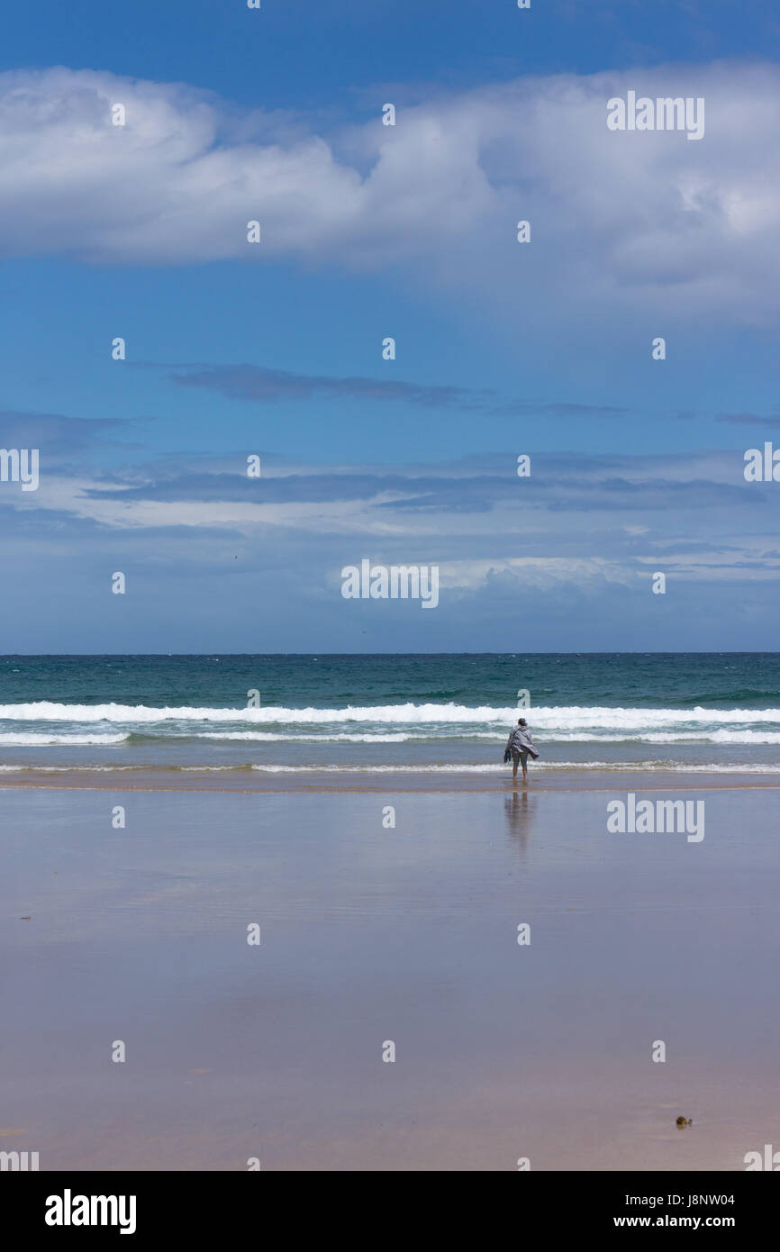 Una donna sta guardando fuori per le onde che si infrangono sulla riva con una leggera brezza su un inizio di giornata di primavera, con il cielo blu e nuvole di luce. Foto Stock