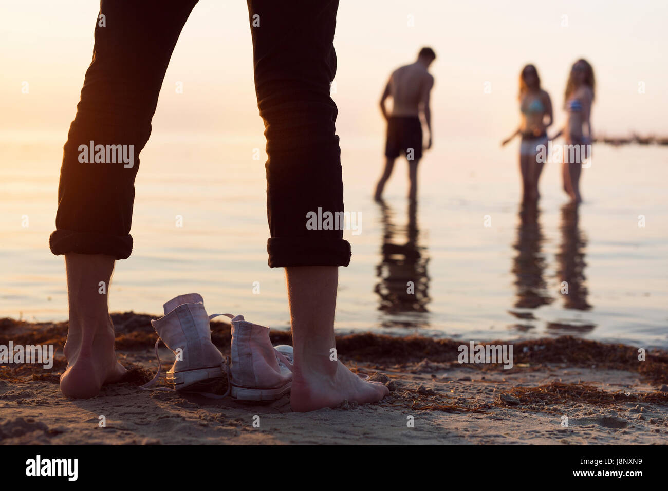 Giovane uomo in piedi sulla spiaggia mentre giovane donna e ragazza adolescente (16-17) e il giovane uomo guadare in mare al tramonto Foto Stock