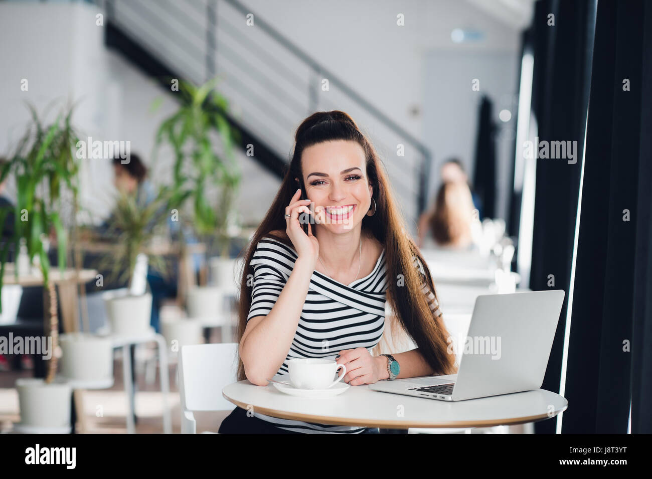 Bella donna adulta con bel sorriso sta parlando al telefono e di lavorare con un computer portatile in un bar mentre guardando lontano. Foto Stock