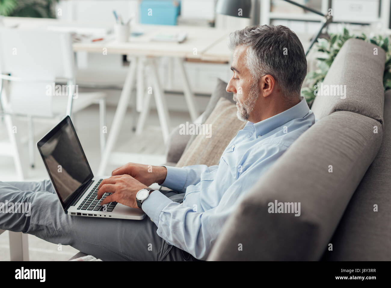 Imprenditore a casa seduti sul divano con i piedi e di lavoro con il suo computer portatile Foto Stock