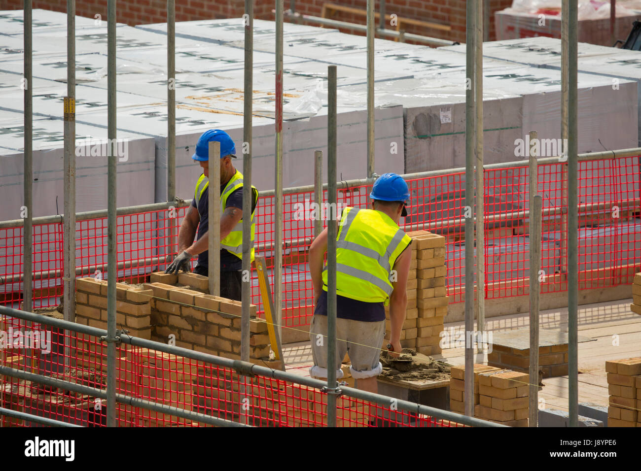 Due muratori o lavoratori edili posando mattoni su un nuovo alloggiamento greenfield lo sviluppo con i ponteggi e la rete di sicurezza Foto Stock