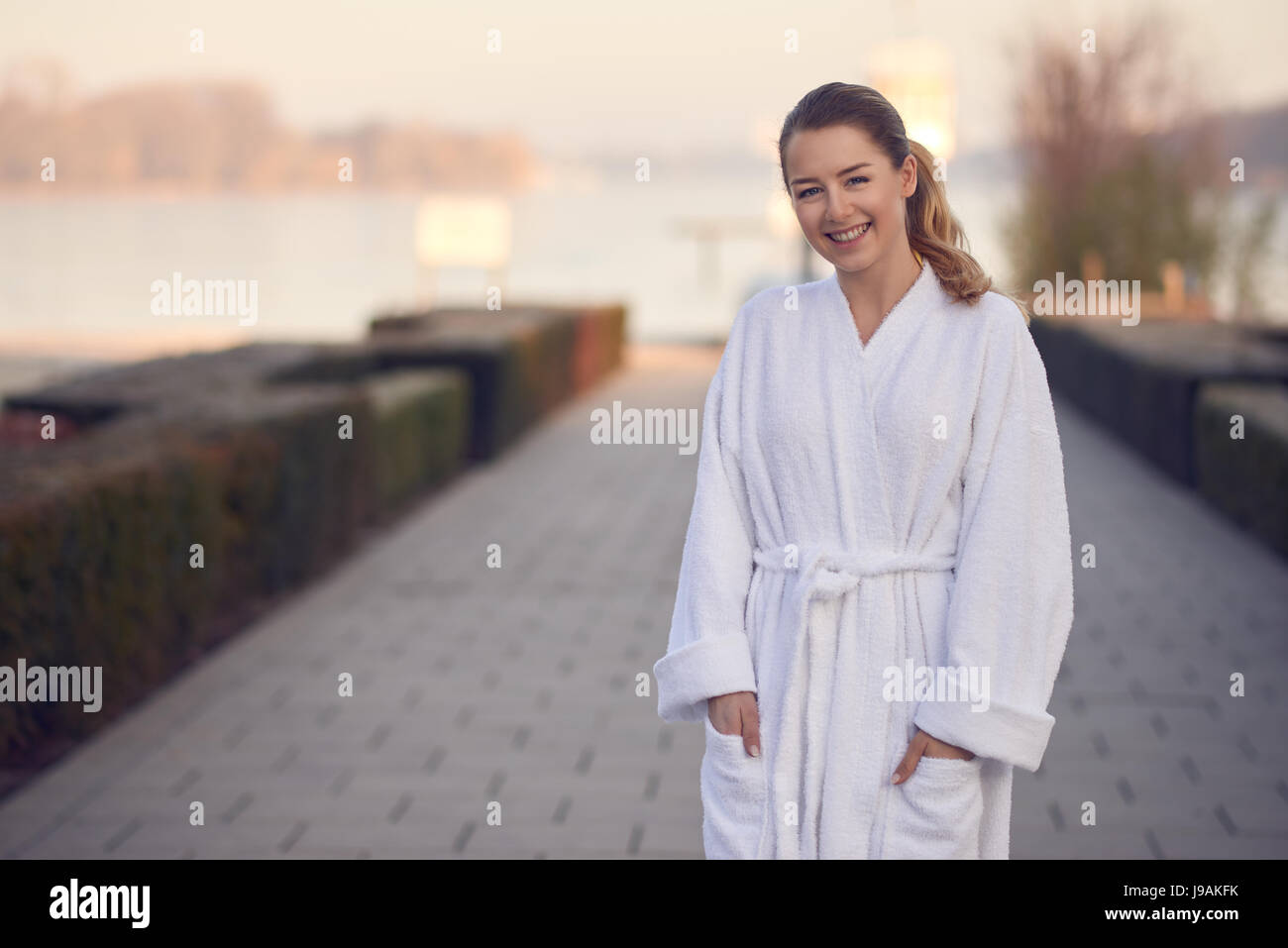 Sorridente giovane donna in un accappatoio bianco in piedi all'aperto su di un viale lastricato che conduce a un tranquillo lago con le sue mani in tasca in un centro benessere un Foto Stock