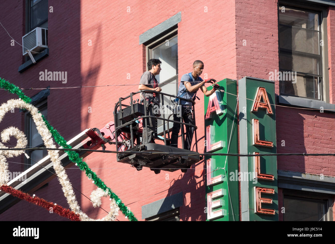 Due giovani uomini al lavoro da un sollevamento platformed per attaccare un segno elettrico attraverso la larghezza del Mulberry Street in Little Italy, NYC Foto Stock