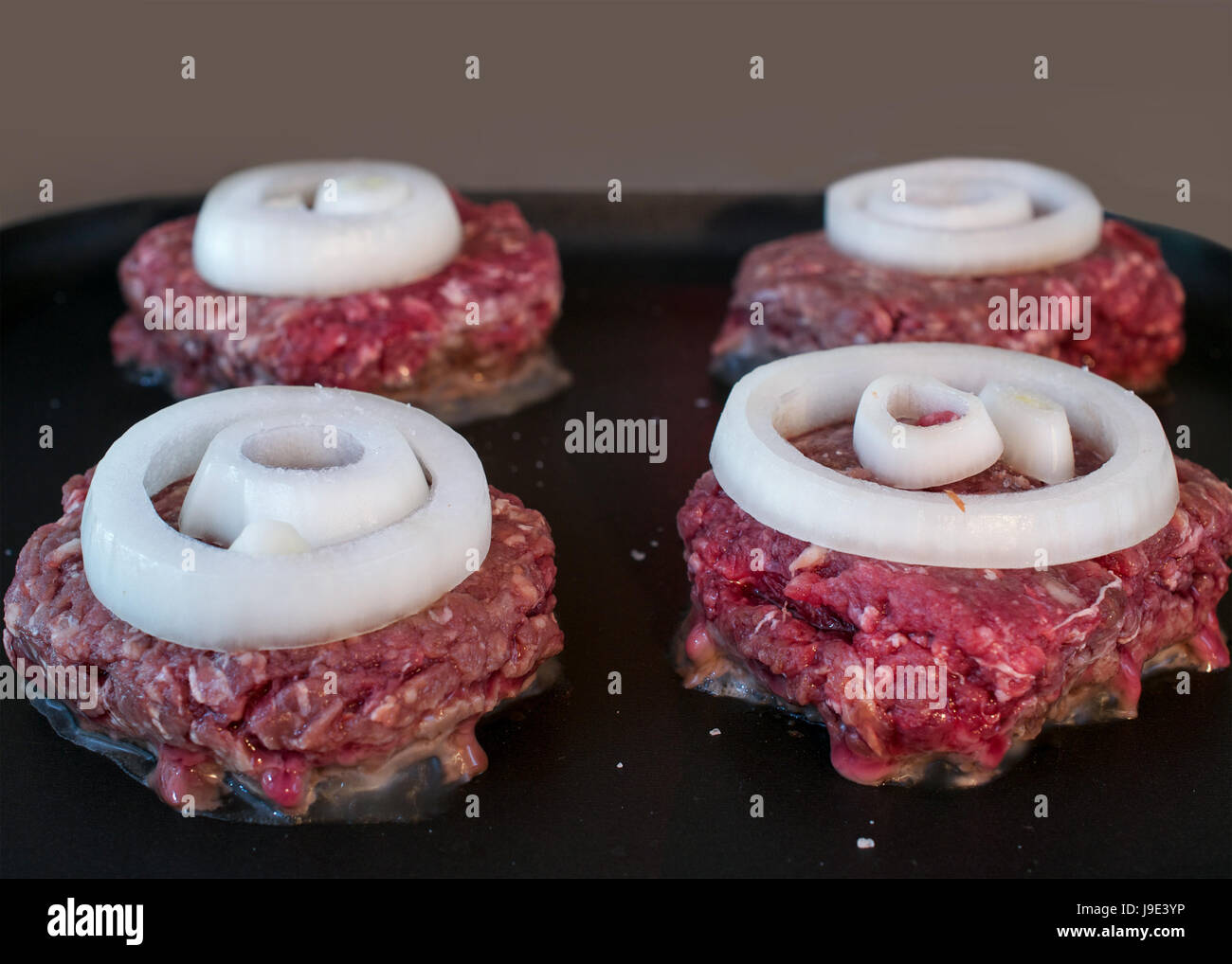 La cottura di quattro materie hamburger di carne di cervo guarnito con anelli di cipolla in padella antiaderente sulla parte superiore di una stufa elettrica, primo piano Foto Stock