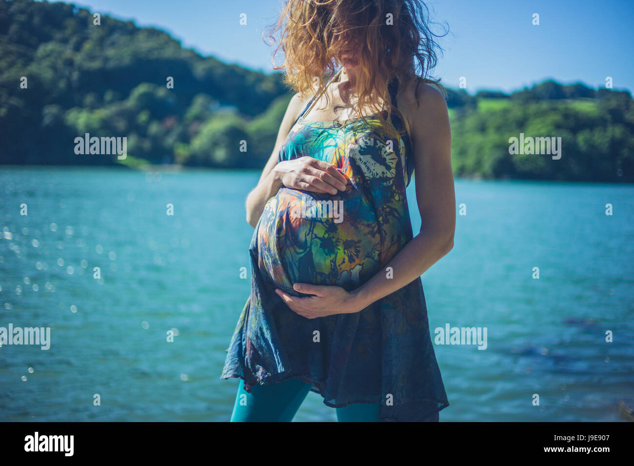 Una giovane donna incinta è in piedi vicino al fiume in una foresta in una giornata di sole di estate Foto Stock