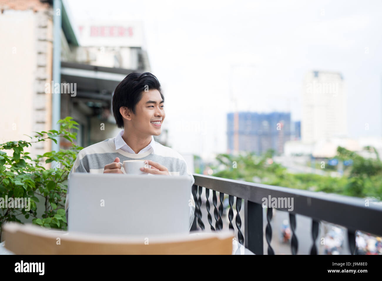 Bello asian giovane uomo che lavora sul computer portatile e sorridere mentre godendo il caffè guardando fuori alla vista Foto Stock