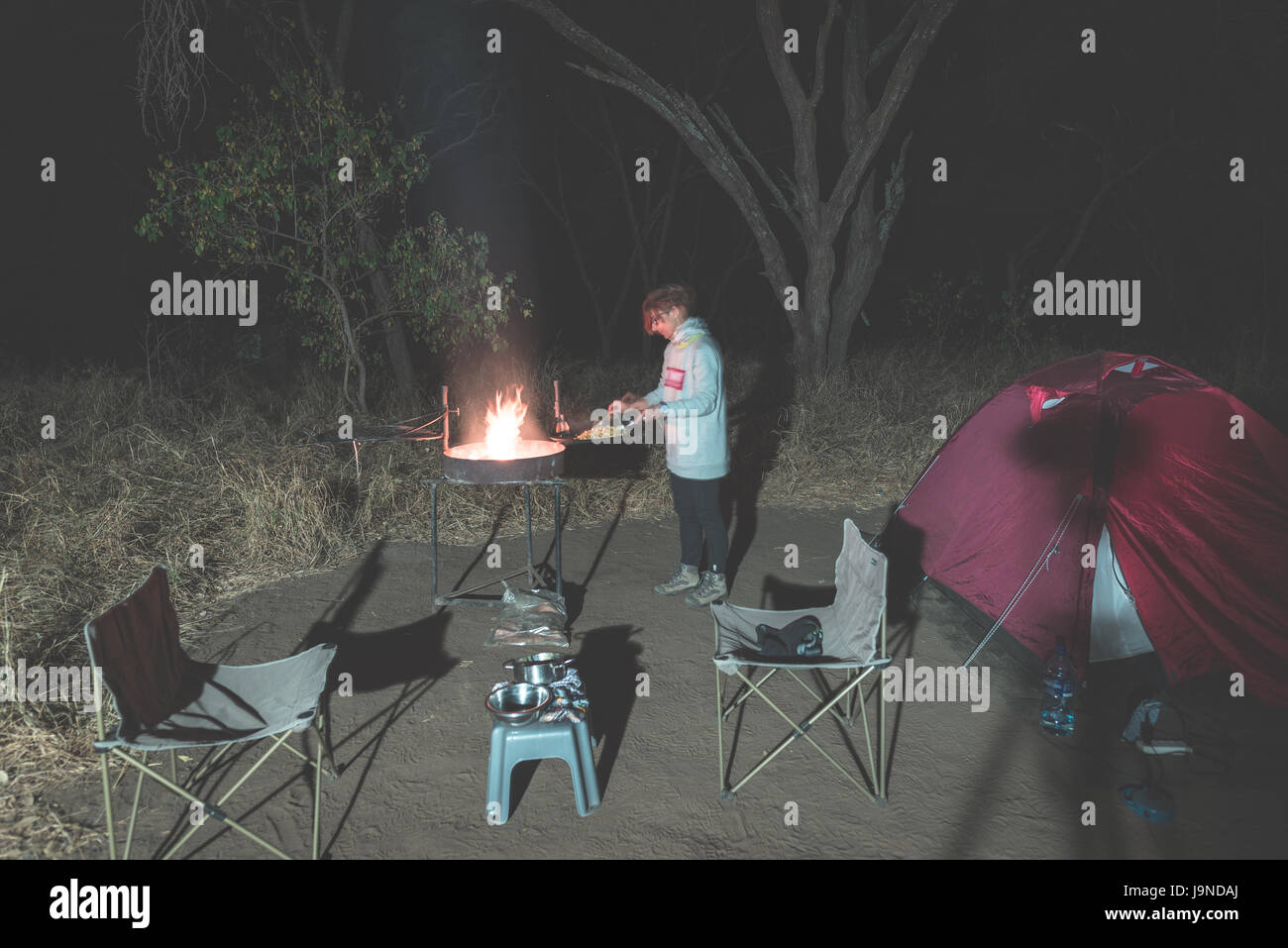 La donna la cottura con fuoco a legna e attrezzature braai di notte. Tenda e sedie in primo piano. Avventure in Africa parchi nazionali. Tonica immagine. Foto Stock