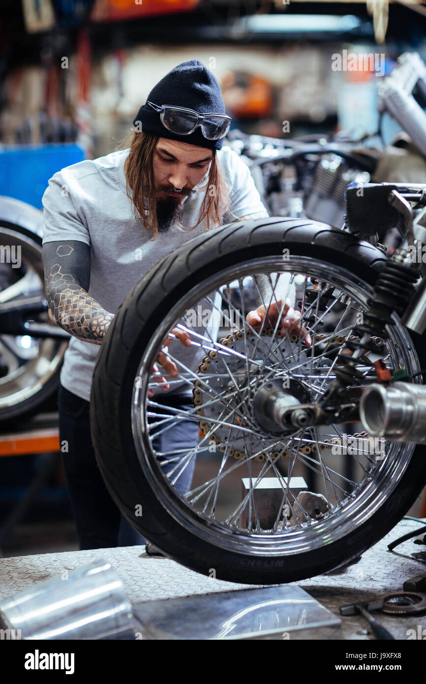Ritratto della moderna tatuato uomo che lavora in garage personalizzazione delle moto e riparazione rotto il meccanismo della ruota Foto Stock