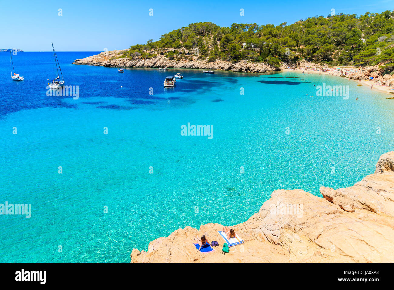 Unidentified due giovani donne a prendere il sole su una roccia a Cala Salada bay con belle crystal clear acqua del mare turchese, isola di Ibiza, Spagna Foto Stock