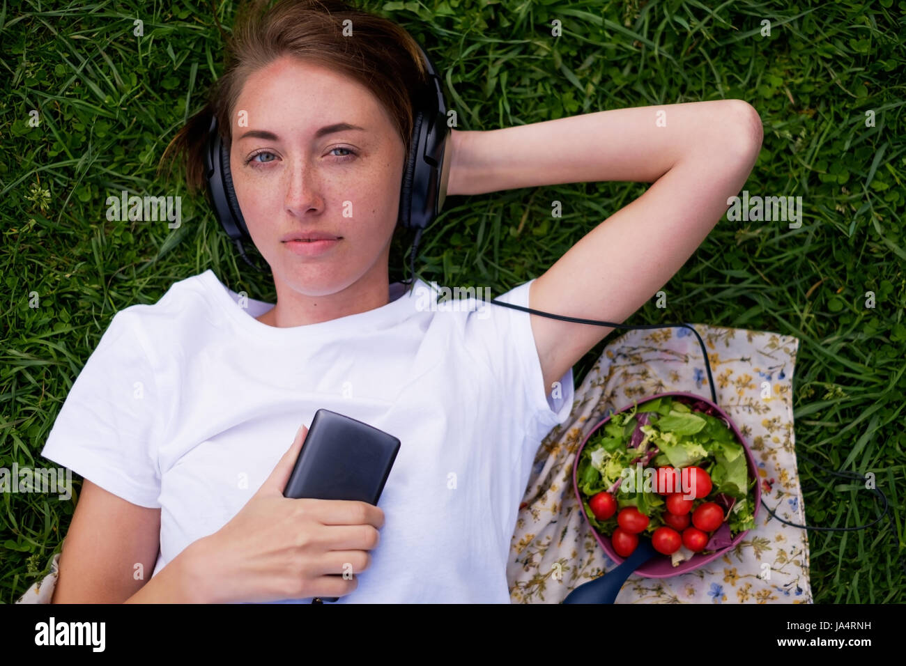 Una bella ragazza nelle cuffie si trova sull'erba verde nel parco e ama ascoltare la musica con piacere. Avere un periodo di riposo nella natura d'estate. N Foto Stock