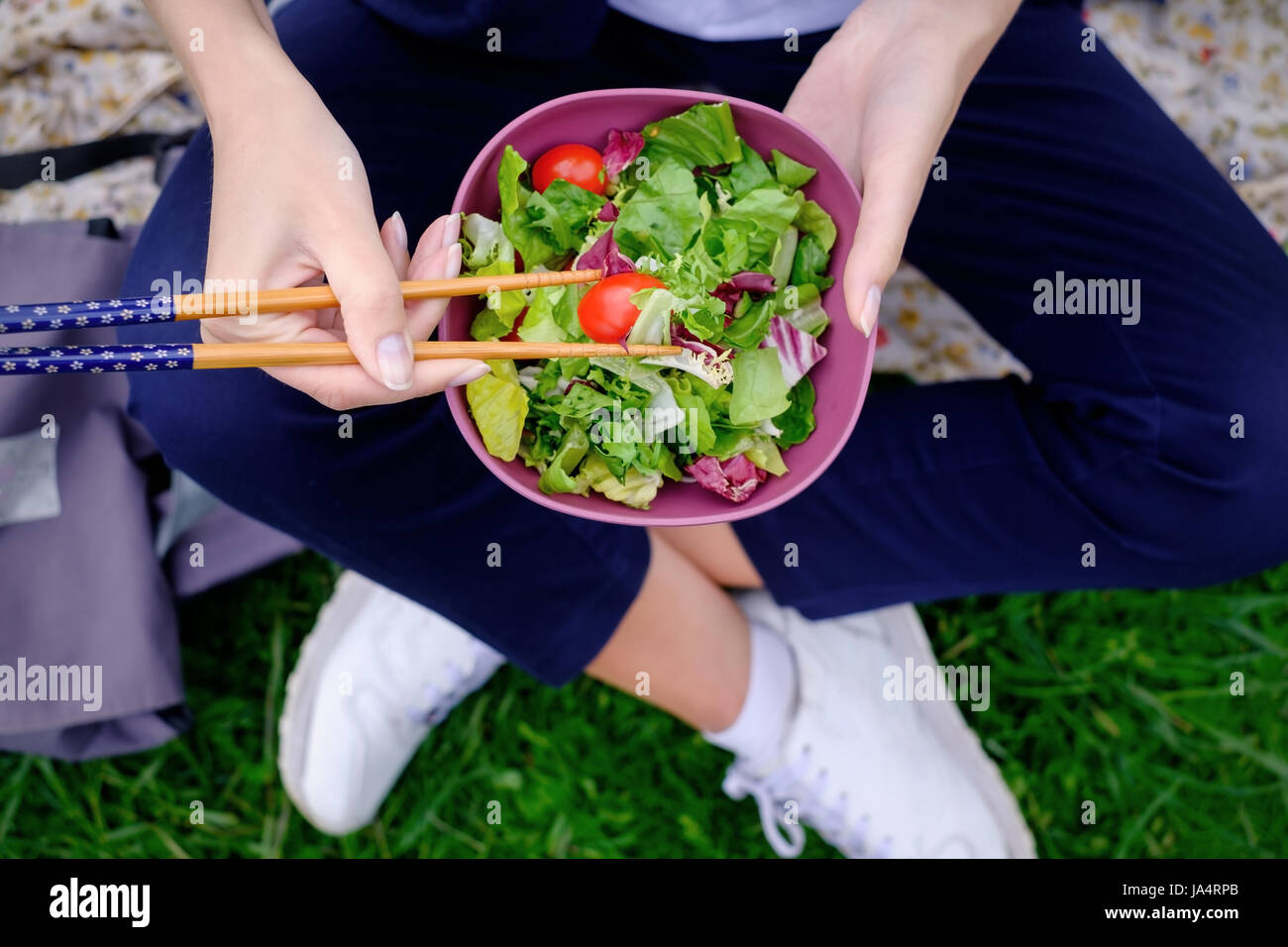 Una ragazza si siede sul prato e mangia un verde insalata fresca con bacchette per sushi. Una sana dieta su alimenti vegetali. Foto Stock