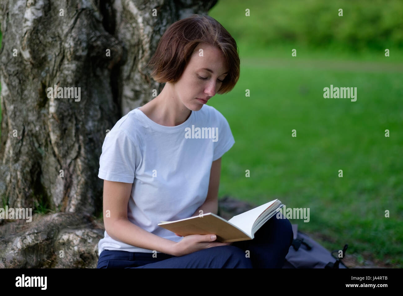 Una ragazza legge un libro in un parco seduto da un albero sul prato verde. Rilassatevi all'aperto estate Foto Stock