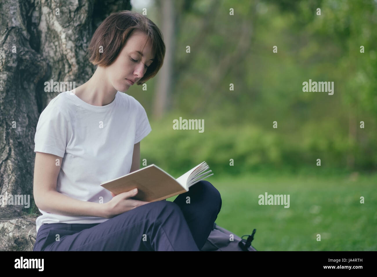Una ragazza legge un libro in un parco seduto da un albero sul prato verde. Rilassatevi all'aperto estate Foto Stock