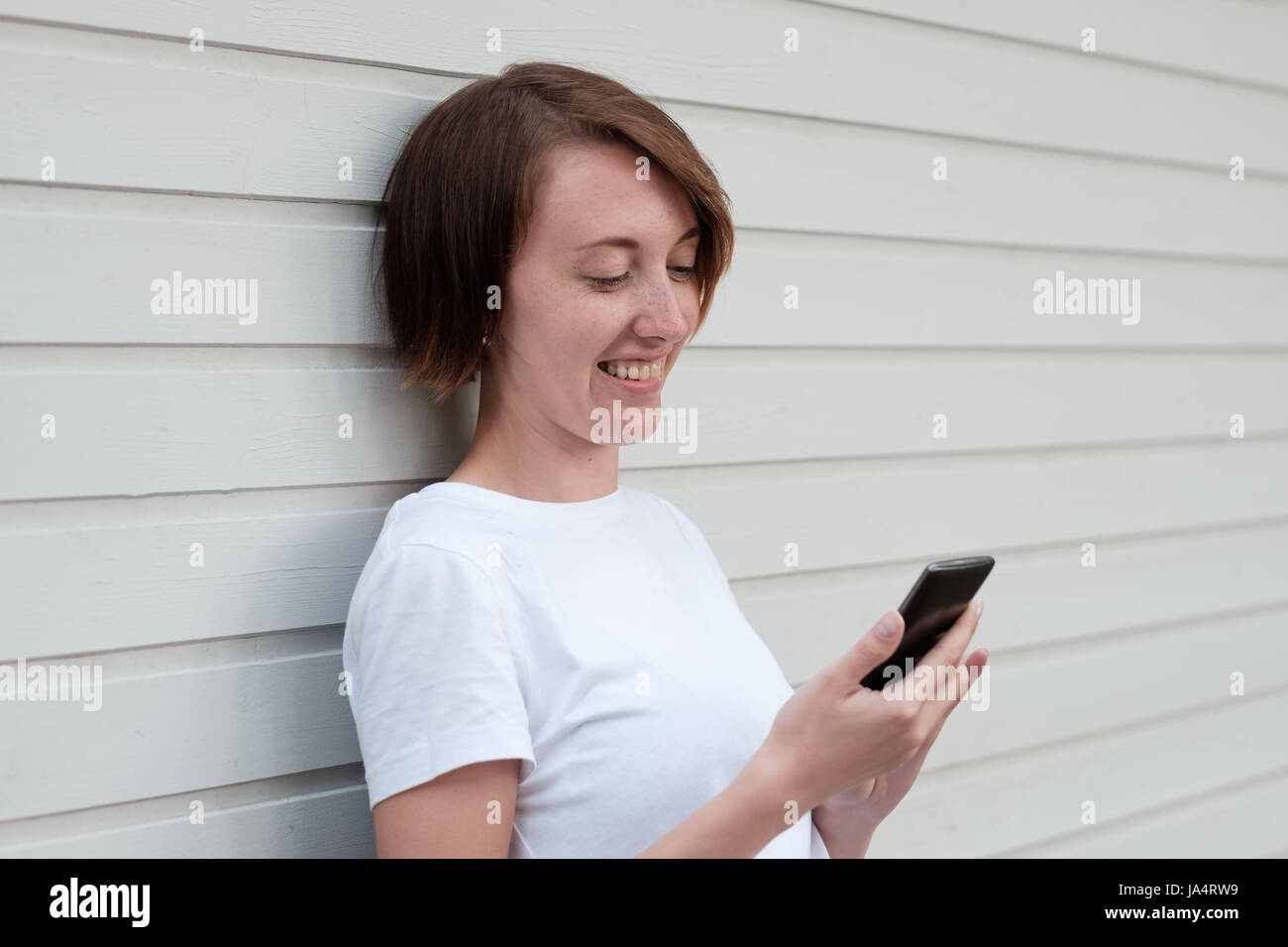 Una ragazza con le lentiggini è ai piedi della parete in legno e lettura sms nel telefono. La comunicazione attraverso i moderni dispositivi. Foto Stock