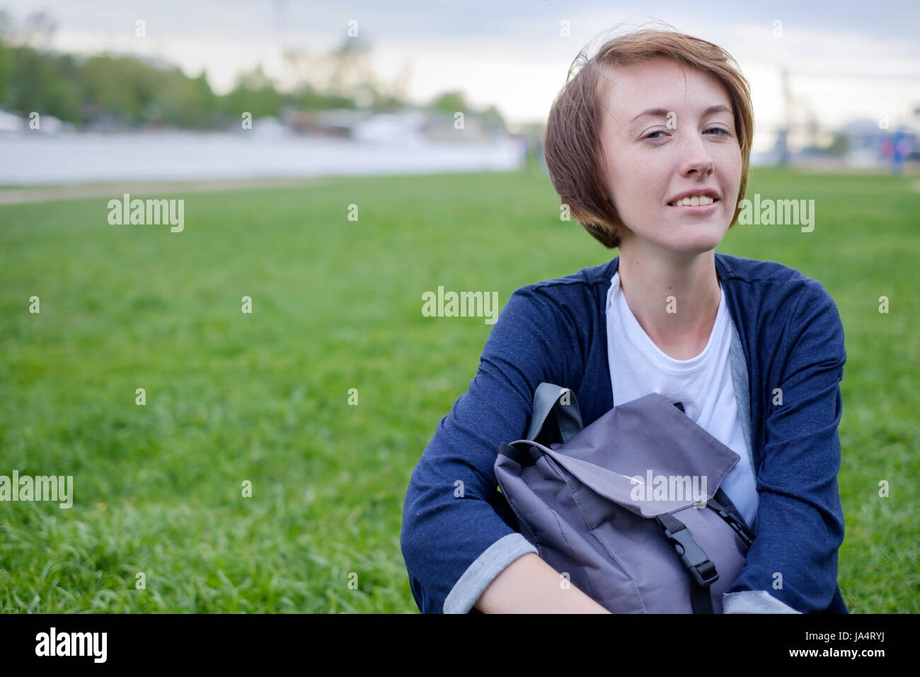 Una ragazza con le lentiggini si siede sul prato verde nel parco. Lei si mantiene uno zaino nelle sue mani. Avere un periodo di riposo in estate all'aria fresca. Foto Stock